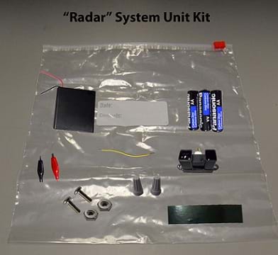 Photo shows a clear gallon-sized plastic zip-lock bag with battery holder, batteries, sensor, alligator clips, two screws, two hex nuts, and two wire connectors. The image is labeled '"Radar" System Unit Kit.