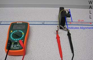 Photo shows a multimeter, measuring tape, and box to calibrate a Sharp GP2Y0A02YK0F Sensor. The sensor is facing the wall is connected to the multimeter by two wires, one red and one black with alligator clips. 