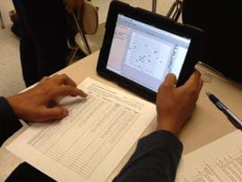 The photo shows a student at a table with a tablet computer showing a graph with a paper listing of data nearby.
