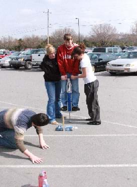 A photograph shows four teens in a parking lot, one on a pogo stick, two helping the boy on the pogo stick and one on his knees, looking at the lower part of the pogo stick.