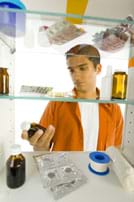 Photo shows young man looking into a medicine cabinet and holding a brown bottle of liquid medicine. 