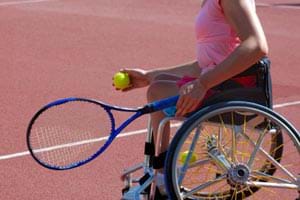 A woman with a physical disability sitting in a wheelchair holding a tennis raquet and tennis ball.
