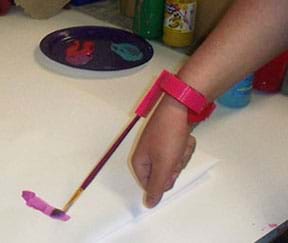 A photograph shows a person’s forearm painting on a piece of paper by the use of an assistive device holds a small paintbrush and is attached to her wrist.