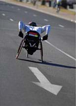 Photo shows a competitor in a sporty wheelchair racing up an asphalt road.