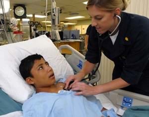 A female nurse checks on a young male patient in a hospital bed by holding a stethoscope on his chest.