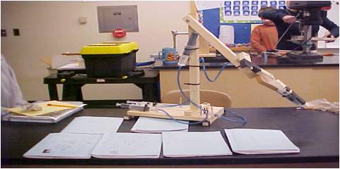 Photo shows a hydraulic arm made of wood on a desk with several engineering design journals.