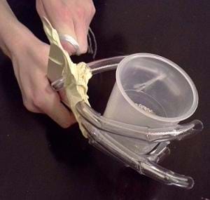 A photograph shows two hands manipulating a student-created prototype made of clear plastic tubing, tape and craft sticks. The prototype is gripping a plastic cup.