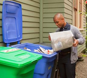 A photograph shows a man emptying a wastebasket of paper into a bigger blue recycling bin full of paper.