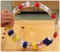 A photograph of a sensory toy shows a ring made of clear plastic tubing covered with scattered stripes of tape in bright colors with moving bells and balls inside the tube.