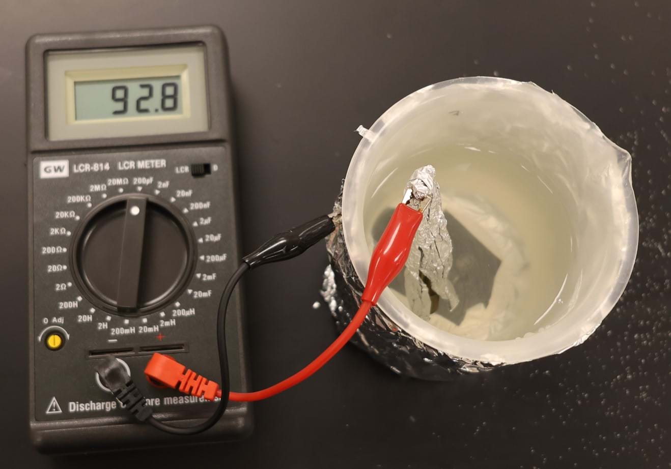 An LCR meter has a black and a red clip attached to it. These clips are attached to the two aluminum sheets used in the experiment. The black clip is hooked to the sheet that is wrapped around the cylinder, while the red clip is attached to the foil that is in the saltwater solution. An LCR meter has a black and a red clip attached to it. These clips are attached to the two aluminum sheets used in the experiment. The black clip is hooked to the sheet that is wrapped around the cylinder, while the red clip is attached to the foil that is in the saltwater solution.