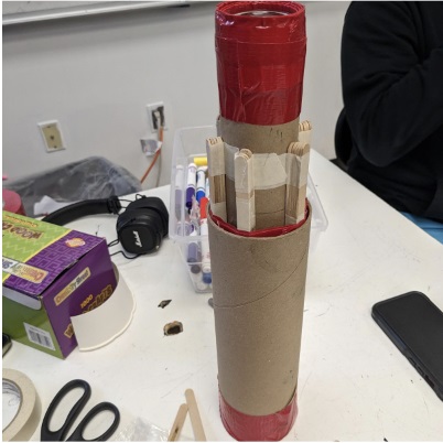 A photo showing a circular tube on a white table facing upright, consisting of two parts, with the inner part, including popsicle sticks, on the outside. There is one lens on each side, with the lens supported by red tape.