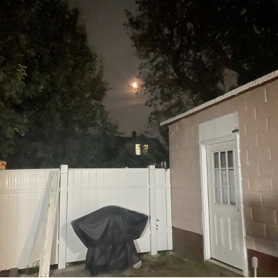 A photo showing a night-time image of the moon in the far distance covered by clouds, with a white fence and light pink side of a house in the foreground.