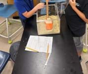 A photo showing two students dropping a fishing weight into a square container of kinetic sand. One student is holding a meter stick next to the container. A photo showing two students dropping a fishing weight into a square container of kinetic sand. One student is holding a meter stick next to the container.