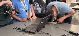 A photo showing a group of students standing around a table in a classroom. One student is leaning over and looking closely at a laptop screen while others observe. Various cables and small electronic components are connected to the laptop. A photo showing a group of students standing around a table in a classroom. One student is leaning over and looking closely at a laptop screen while others observe. Various cables and small electronic components are connected to the laptop.