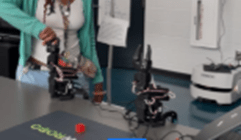 A photo showing a student standing at a table in a classroom, adjusting a small robotic arm connected by wires. Another robotic arm sits nearby on the table. A laptop and additional equipment are also visible. A photo showing a student standing at a table in a classroom, adjusting a small robotic arm connected by wires. Another robotic arm sits nearby on the table. A laptop and additional equipment are also visible.