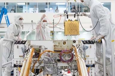 A photo showing three engineers wearing white protective cleanroom suits, gloves, and face masks working inside a laboratory to install a gold, cube-shaped instrument into the open body of a Mars rover. The rover is surrounded by exposed wiring, tubing, and mechanical components, with solar panels visible on either side. A photo showing three engineers wearing white protective cleanroom suits, gloves, and face masks working inside a laboratory to install a gold, cube-shaped instrument into the open body of a Mars rover. The rover is surrounded by exposed wiring, tubing, and mechanical components, with solar panels visible on either side.