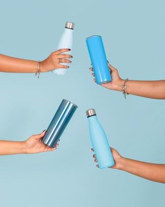 A photo showing four hands with diverse skin tones reaching in from the left and right sides of the frame against a solid pale blue background. Each hand holds a different reusable water bottle or tumbler in varying shades of blue. A photo showing four hands with diverse skin tones reaching in from the left and right sides of the frame against a solid pale blue background. Each hand holds a different reusable water bottle or tumbler in varying shades of blue.