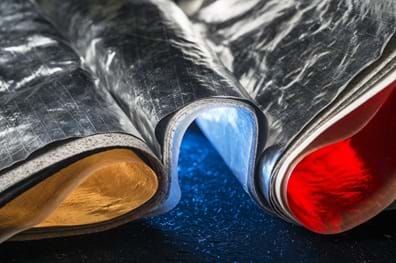 A photo showing a close-up view of layered thermal insulation material. The outer layer is a crinkled, reflective silver foil. The textures appear metallic and industrial, set against a dark, speckled base. A photo showing a close-up view of layered thermal insulation material. The outer layer is a crinkled, reflective silver foil. The textures appear metallic and industrial, set against a dark, speckled base.