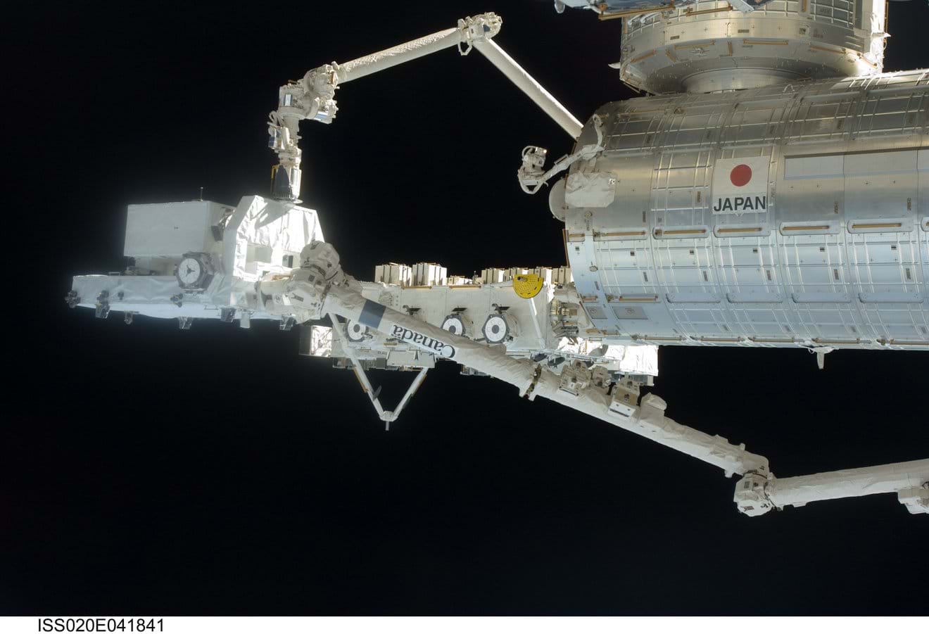 A photo showing a view of the International Space Station modules against the blackness of space. In the foreground, the Japanese Experiment Module (JEM), Kibo, is visible, labeled with the word 'JAPAN'. Attached to the module are two highly articulated robotic arms. The larger arm, which appears to be the JEM Remote Manipulator System (JEMRMS), extends horizontally, and a smaller, more delicate arm is positioned above it, showing the complex capabilities required for station assembly and maintenance. A photo showing a view of the International Space Station modules against the blackness of space. In the foreground, the Japanese Experiment Module (JEM), Kibo, is visible, labeled with the word 'JAPAN'. Attached to the module are two highly articulated robotic arms. The larger arm, which appears to be the JEM Remote Manipulator System (JEMRMS), extends horizontally, and a smaller, more delicate arm is positioned above it, showing the complex capabilities required for station assembly and maintenance.