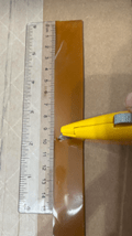 A photo showing a close-up of a rectangular strip of orange-brown material placed next to a transparent plastic ruler. The ruler is marked in centimeters, with the material aligned along its length for measurement. A yellow box cutter is positioned at the edge of the material, with the blade extended, indicating that the material is in the process of being cut at 10 cm. 