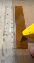 A photo showing a close-up of a transparent plastic ruler placed next to a rectangular strip of orange-brown material. A yellow box cutter is being used to cut a slit in the material at 9 cm.