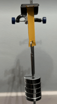 A photo showing a stress testing setup. A strip of material is clamped at the top of a lab stand. The strip is vertically suspended, and a set of cylindrical metal weights is attached to the bottom of the strip, applying downward force. 