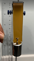 A photo showing a stress testing setup. A strip of material is clamped at the top of a lab stand. The strip is vertically suspended, and a set of cylindrical metal weights is attached to the bottom of the strip, applying downward force. A clear plastic ruler is lined next to the strip showing the strip length to be 10 cm.