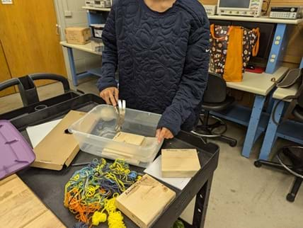 A photo showing a student engaged in a tap testing activity as part of a non-destructive evaluation (NDE) exercise in a classroom or laboratory setting. The student is wearing a dark, quilted sweatshirt and is using a hammer to tap a wooden block.