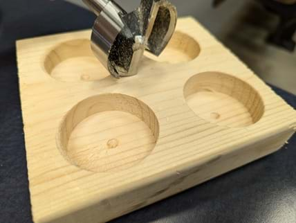 An image showing a close-up view of a Forstner bit resting on a wooden block. The wooden block measures approximately 6 inches by 2 inches by 6 inches. The block has four cylindrical holes drilled into it, each positioned in one of the four quadrants of the top surface. The holes are evenly spaced and appear smooth and well-defined, indicative of the clean cutting action of the Forstner bit. The Forstner bit itself, metallic and slightly worn from use, is positioned above one of the holes, laying on its side with the sharp edges visible.