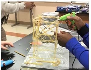 A photo showing a student using a hot glue gun to make a connection between spaghetti pieces as they construct their building. 