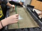 A photo showing students using a straight edge to smooth the surface of the sand in the stream table.