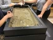 A photo showing students around a stream table with a funnel positioned to increase the water flow.