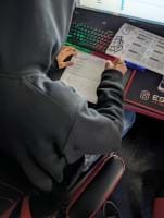 A photo showing a student wearing a dark hoodie seated at a computer station with a red-and-black esports-themed desk. The student is working on a printed worksheet titled "4 Input Logic Gate," using a pencil to complete a truth table, referencing a logic gates handout while looking at a screen that displays gate logic information. A photo showing a student wearing a dark hoodie seated at a computer station with a red-and-black esports-themed desk. The student is working on a printed worksheet titled "4 Input Logic Gate," using a pencil to complete a truth table, referencing a logic gates handout while looking at a screen that displays gate logic information.