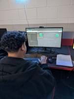 A photo of a student sitting at a computer desk using Wokwi, a logic circuit simulator, to test a custom logic gate design. The student is wearing a dark hoodie and is focused on the monitor, which displays logic gates, input switches, and a code window. A worksheet labeled “Logic Gates III” sits on the desk, partially completed with drawn gates and truth table outputs. A photo of a student sitting at a computer desk using Wokwi, a logic circuit simulator, to test a custom logic gate design. The student is wearing a dark hoodie and is focused on the monitor, which displays logic gates, input switches, and a code window. A worksheet labeled “Logic Gates III” sits on the desk, partially completed with drawn gates and truth table outputs.