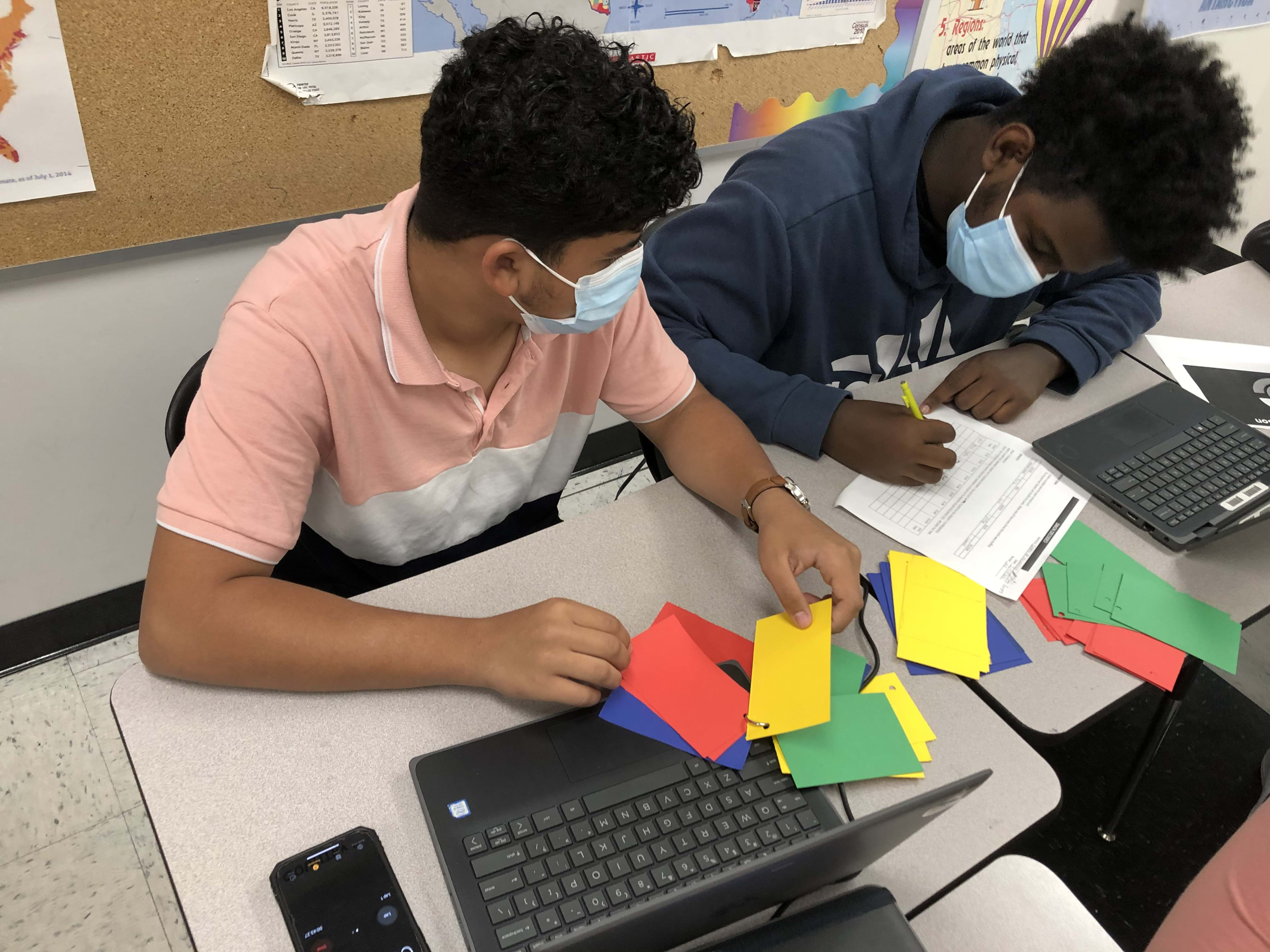 A photo of two students playing an analog “Simon” game with colored index cards; one is looking through the colored cards and the other is writing on the activity worksheet.