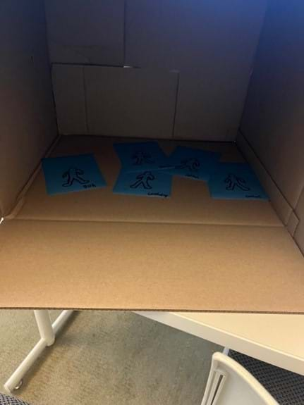 A photo showing five sheets of blue photoluminescent paper inside a large box. Each sheet has a person-shape drawn on it, and each sheet is labeled with the fabric to be tested or as the control.