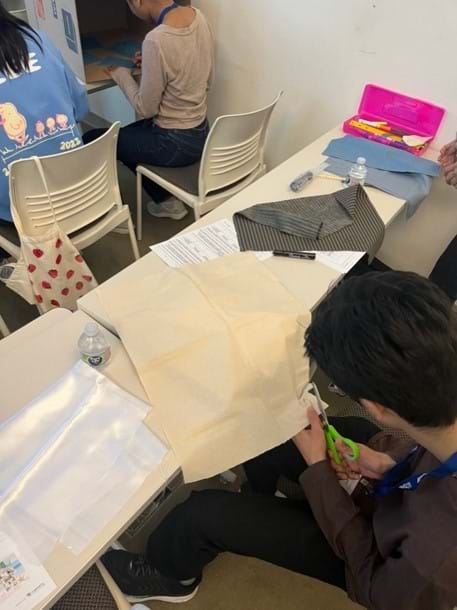 A photo of a student cutting a piece of beige cotton fabric using scissors. 