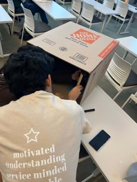 A photo showing a student reaching inside their box to measure the distance between the UV flashlight and their person-shape on the photoluminescent paper with a ruler.