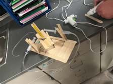 A photo showing a flat base constructed of wide popsicle sticks with a short popsicle stick tower in the center. On the tower are 4 wide popsicles put together as a platform that can rotate as the syringe pushes against it. The syringes are attached to the platform using popsicle sticks. When you push one, it causes the other syringe to extend out, pushing and moving the platform. A photo showing a flat base constructed of wide popsicle sticks with a short popsicle stick tower in the center. On the tower are 4 wide popsicles put together as a platform that can rotate as the syringe pushes against it. The syringes are attached to the platform using popsicle sticks. When you push one, it causes the other syringe to extend out, pushing and moving the platform.