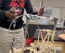 A photo showing a student pushing 2 syringes, one in each hand, attached to a plastic tube with a horizontal syringe on each side. The horizontal syringes are attached using duct tape to a thin skewer that has a small cube on it. Attached to the cube is a thin string holding a small platform made from small popsicle sticks. The skewer holding all of this is attached to a rectangular base made from popsicle sticks, with 2 longer popsicle stick towers (held together by duct tape) standing above the bottom base. As the student pushed one syringe, the syringe at the other end of the plastic tube extends pushing the block the other direction, and when the student pushes the opposite syringe, the same thing happens but in the other direction. A photo showing a student pushing 2 syringes, one in each hand, attached to a plastic tube with a horizontal syringe on each side. The horizontal syringes are attached using duct tape to a thin skewer that has a small cube on it. Attached to the cube is a thin string holding a small platform made from small popsicle sticks. The skewer holding all of this is attached to a rectangular base made from popsicle sticks, with 2 longer popsicle stick towers (held together by duct tape) standing above the bottom base. As the student pushed one syringe, the syringe at the other end of the plastic tube extends pushing the block the other direction, and when the student pushes the opposite syringe, the same thing happens but in the other direction.