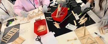 A photo showing three students at a worktable containing popsicles sticks, hot glue guns, syringe packets, plastic tubing, skewers, and other materials. They are constructing parts of their popsicle stick bridges. A photo showing three students at a worktable containing popsicles sticks, hot glue guns, syringe packets, plastic tubing, skewers, and other materials. They are constructing parts of their popsicle stick bridges.