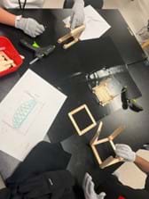 A photo showing two students constructing their bridges using popsicle sticks. In their work area, there is a basket of materials that includes popsicle sticks and glue sticks. They are using hot glue guns and protective gloves. Students also both have their bridge sketches for reference. A photo showing two students constructing their bridges using popsicle sticks. In their work area, there is a basket of materials that includes popsicle sticks and glue sticks. They are using hot glue guns and protective gloves. Students also both have their bridge sketches for reference.