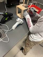 A photo showing a student measuring popsicle sticks glued together with the goal of attaching it to the base of their bridge. The work area is clean, with two hot glue guns and a supply basket visible. The student is also using protective gloves while constructing their bridge. A photo showing a student measuring popsicle sticks glued together with the goal of attaching it to the base of their bridge. The work area is clean, with two hot glue guns and a supply basket visible. The student is also using protective gloves while constructing their bridge.