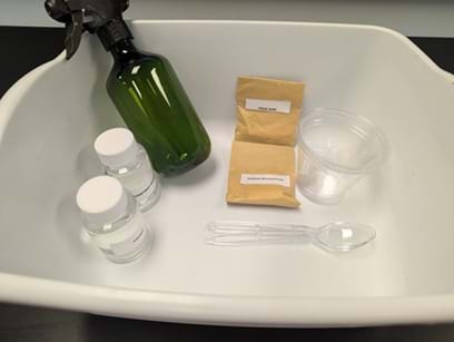 A photo of a white plastic bin containing materials for a hands-on activity. Inside the bin are a green spray bottle, two small clear bottles with white lids, two sealed brown paper packets with labels, a clear plastic cup, a plastic spoon, and two plastic pipettes. The items are arranged loosely inside the bin on a dark tabletop. A photo of a white plastic bin containing materials for a hands-on activity. Inside the bin are a green spray bottle, two small clear bottles with white lids, two sealed brown paper packets with labels, a clear plastic cup, a plastic spoon, and two plastic pipettes. The items are arranged loosely inside the bin on a dark tabletop.