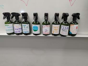A photo of eight dark spray bottles with black trigger sprayers lined up on a whiteboard ledge. Each bottle has a colorful hand-drawn label identifying different cleaning solutions, including names such as “Awesome Dark,” “RA,” “Super Soaker,” “Clean Max,” and “Cool.” The labels use different colors and drawings, indicating the bottles contain different mixtures. The whiteboard behind them has faint marker drawings. A photo of eight dark spray bottles with black trigger sprayers lined up on a whiteboard ledge. Each bottle has a colorful hand-drawn label identifying different cleaning solutions, including names such as “Awesome Dark,” “RA,” “Super Soaker,” “Clean Max,” and “Cool.” The labels use different colors and drawings, indicating the bottles contain different mixtures. The whiteboard behind them has faint marker drawings.