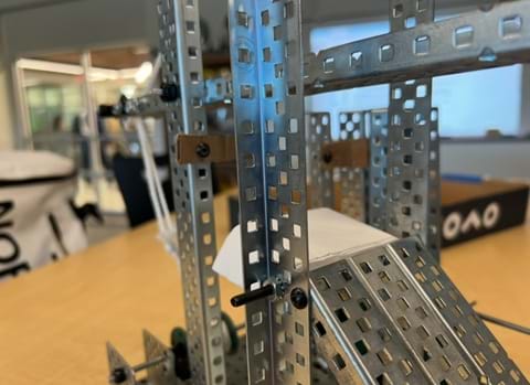 A photo showing a silver steel machine built with VEX parts that has a combination of wheels, gears, levers, and inclined plane. Two parts have been replaced with brown cardboard, and one replaced with white cardstock. A photo showing a silver steel machine built with VEX parts that has a combination of wheels, gears, levers, and inclined plane. Two parts have been replaced with brown cardboard, and one replaced with white cardstock.