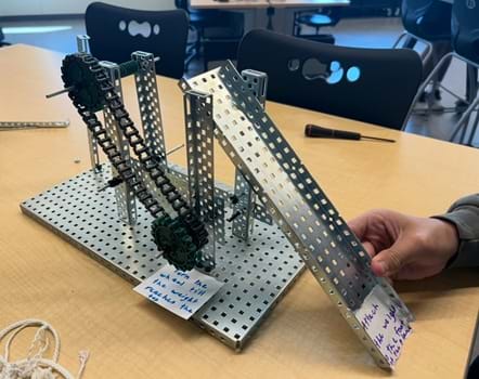 A photo showing a silver steel machine built with VEX parts that has a combination of wheels, gears, levers, and inclined plane. Printed signs are placed at two locations that have dark print on white background to give instructions to end users. A photo showing a silver steel machine built with VEX parts that has a combination of wheels, gears, levers, and inclined plane. Printed signs are placed at two locations that have dark print on white background to give instructions to end users.