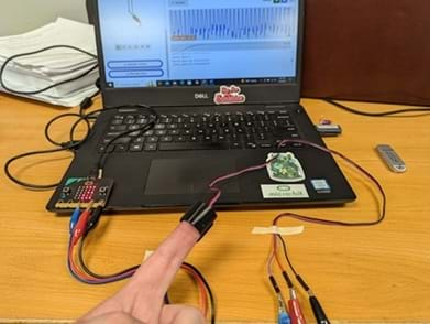 A photo showing a connected micro:bit and pulse sensor recording heart rate. A heartbeat waveform is generated on the screen in the background.  