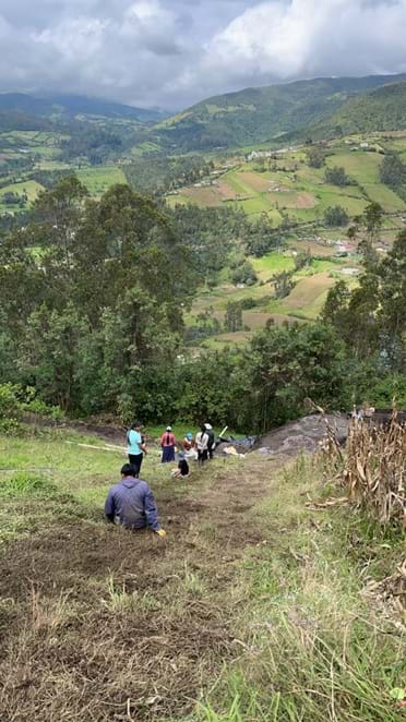 A photo of the same grassy hillside viewed from the top, where people are lined up along a now worn down and muddy path in the grass. A vast agricultural and forested valley near the Yambiro community of Otavalo, Ecuador is in view. A photo of the same grassy hillside viewed from the top, where people are lined up along a now worn down and muddy path in the grass. A vast agricultural and forested valley near the Yambiro community of Otavalo, Ecuador is in view.