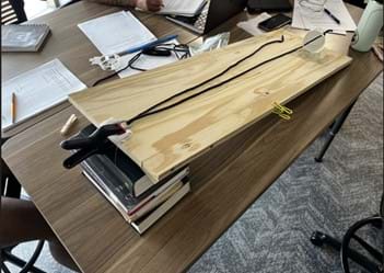 A photo of a student project demonstrating an inclined plane or a similar physics principle using a wooden plank, a stack of books, and a simple pulley system with a string and a white cup. A photo of a student project demonstrating an inclined plane or a similar physics principle using a wooden plank, a stack of books, and a simple pulley system with a string and a white cup.